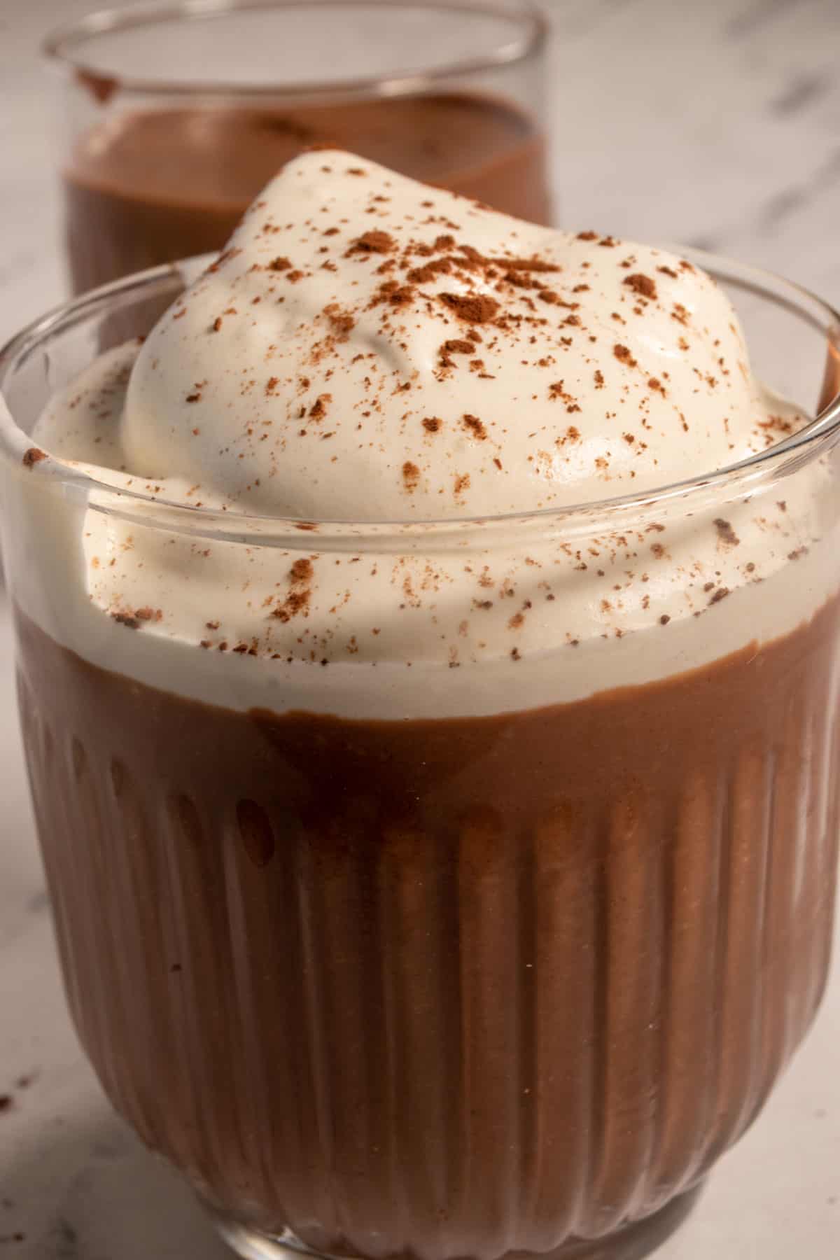 A close up image of chocolate mousse inside a dessert glass. The chocolate inside is thick after setting in the fridge. It is topped with lots of whipped cream.