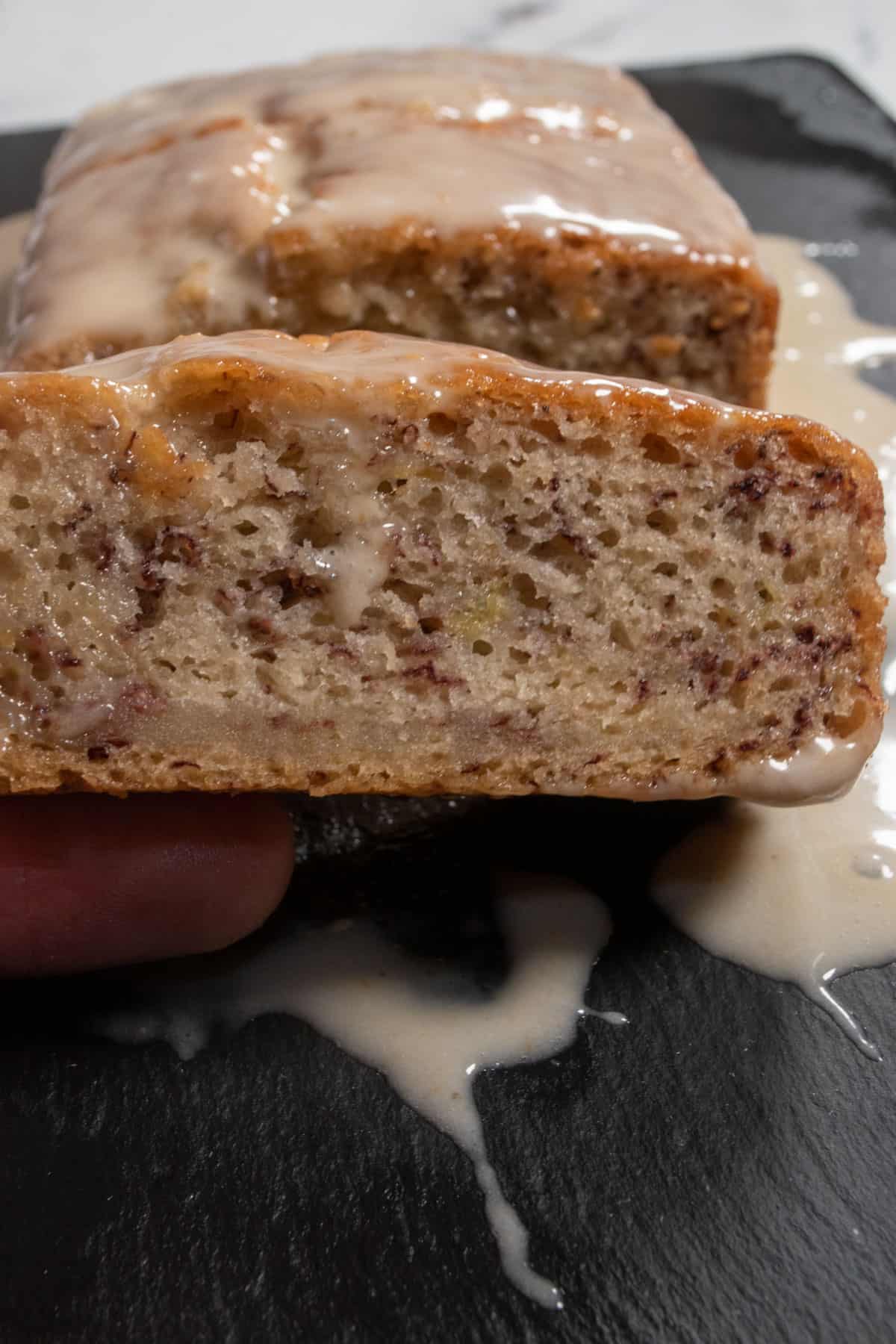A moist, thick slice of banana cake being held by a hand. Glaze has dripped down the side of the cake.