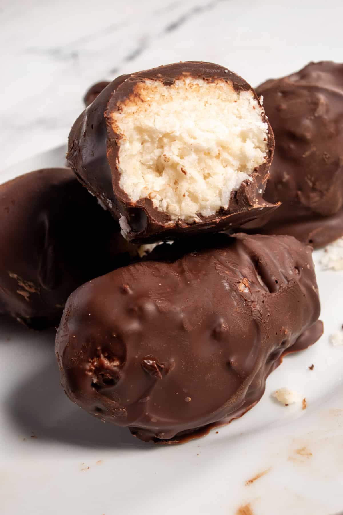 Four vegan bounty bars stacked up on a white platter. The one on top is sliced in half, revealing the coconut filling inside.