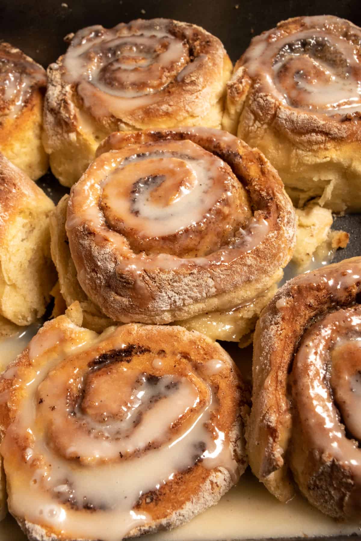 A bunch of golden brown cinnamon rolls in a black pan cooling down. They're all crispy and coated in maple glaze. 