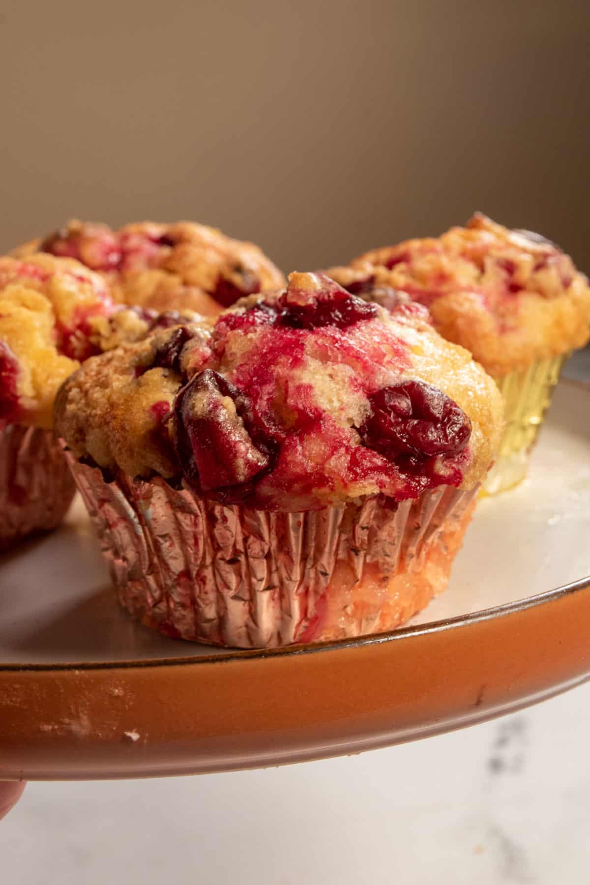 Three vegan cranberry orange muffins on a plate. They have burst making red patterns on top of the muffins.