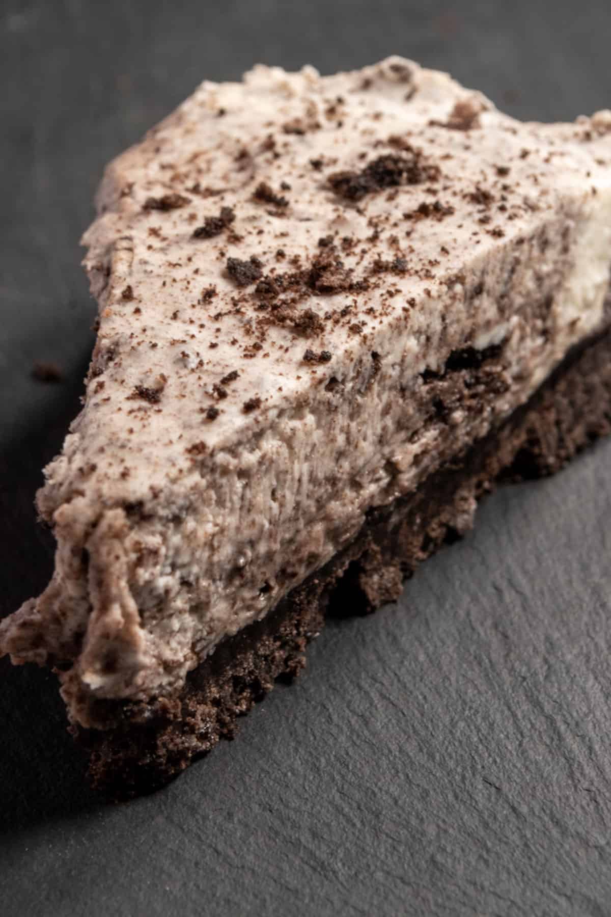 A creamy slice of cheesecake on a slate board. It is topped with extra Oreo crumbs. 