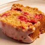 A large slice of cake filled with fresh raspberries and topped with a bright pink raspberry glaze.