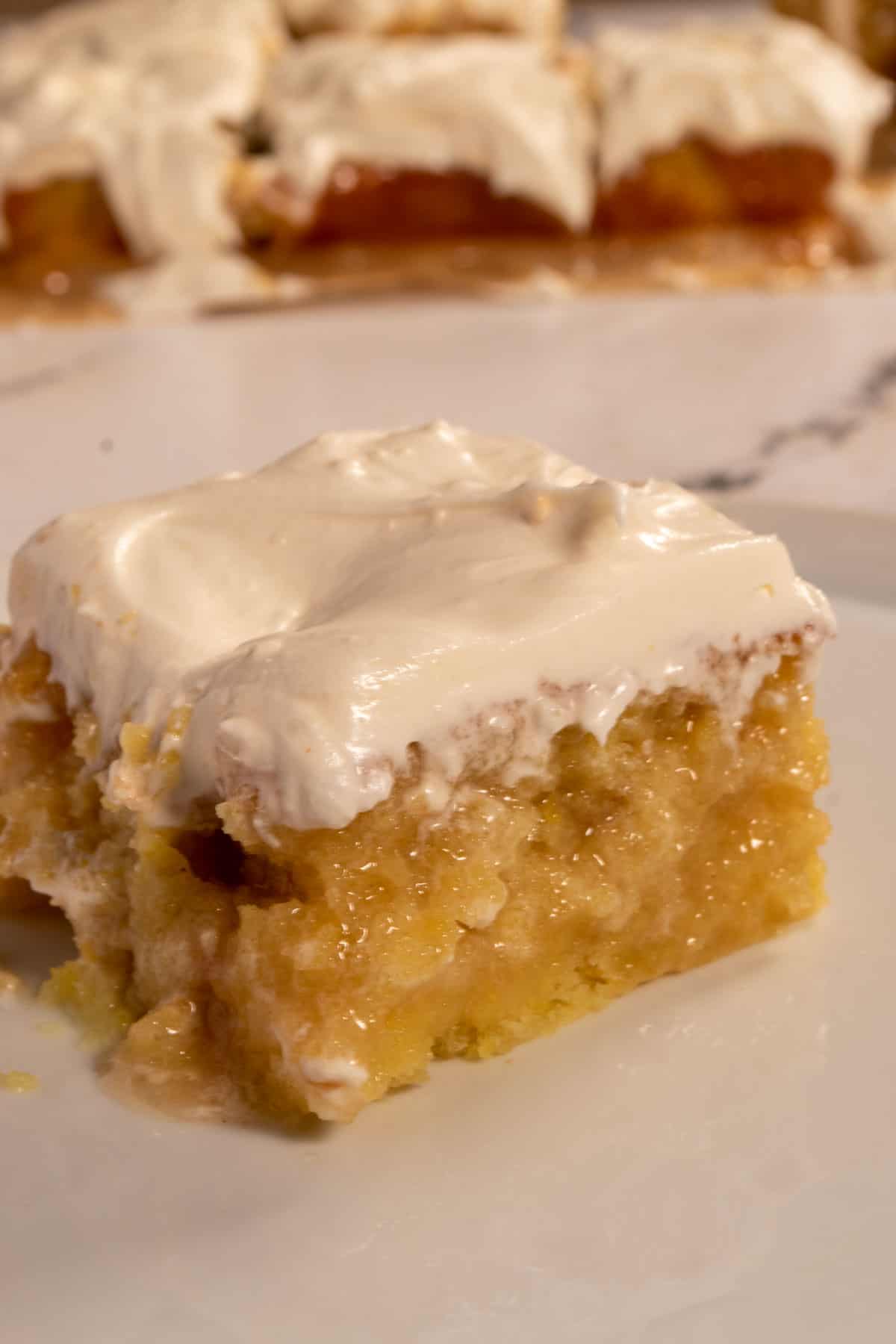 A smaller piece of my moist vegan lemon poke cake on a white plate. Many other slices sit in the background.