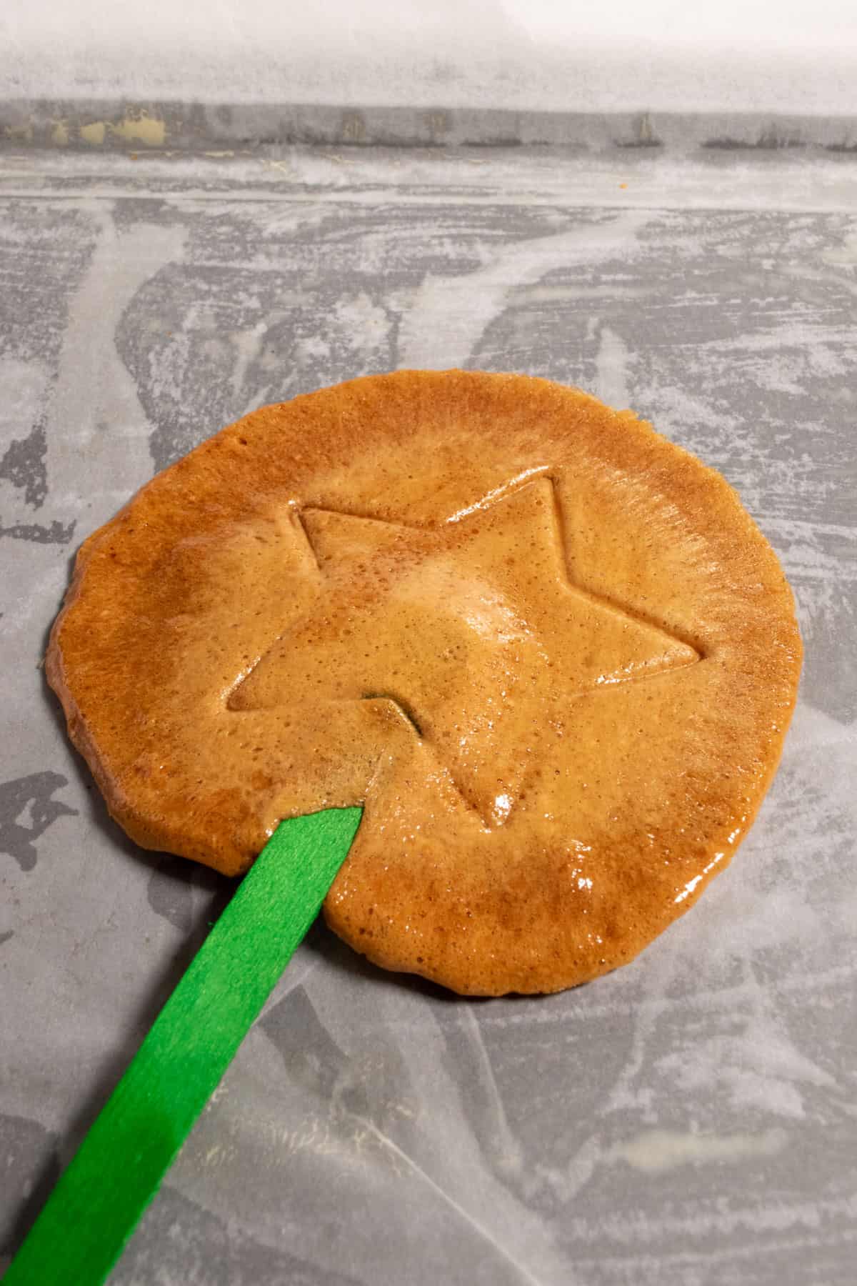 A dalgona laying on a piece of parchment paper. A star has been pressed into the center and it has a bright green popsicle stick pressed into it. 