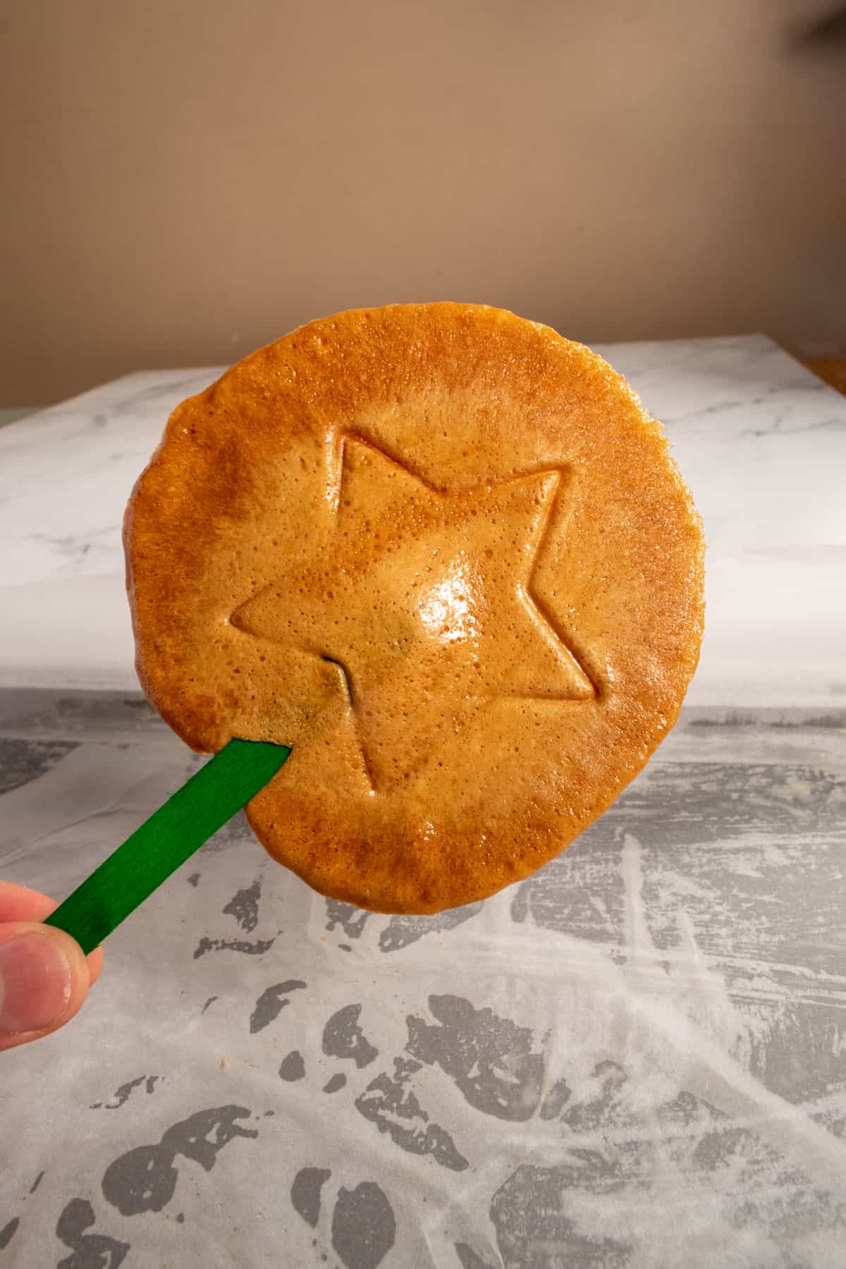 A golden vegan dalgona candy on a green popsicle stick being held by a hand. It has a star imprint in the center. 