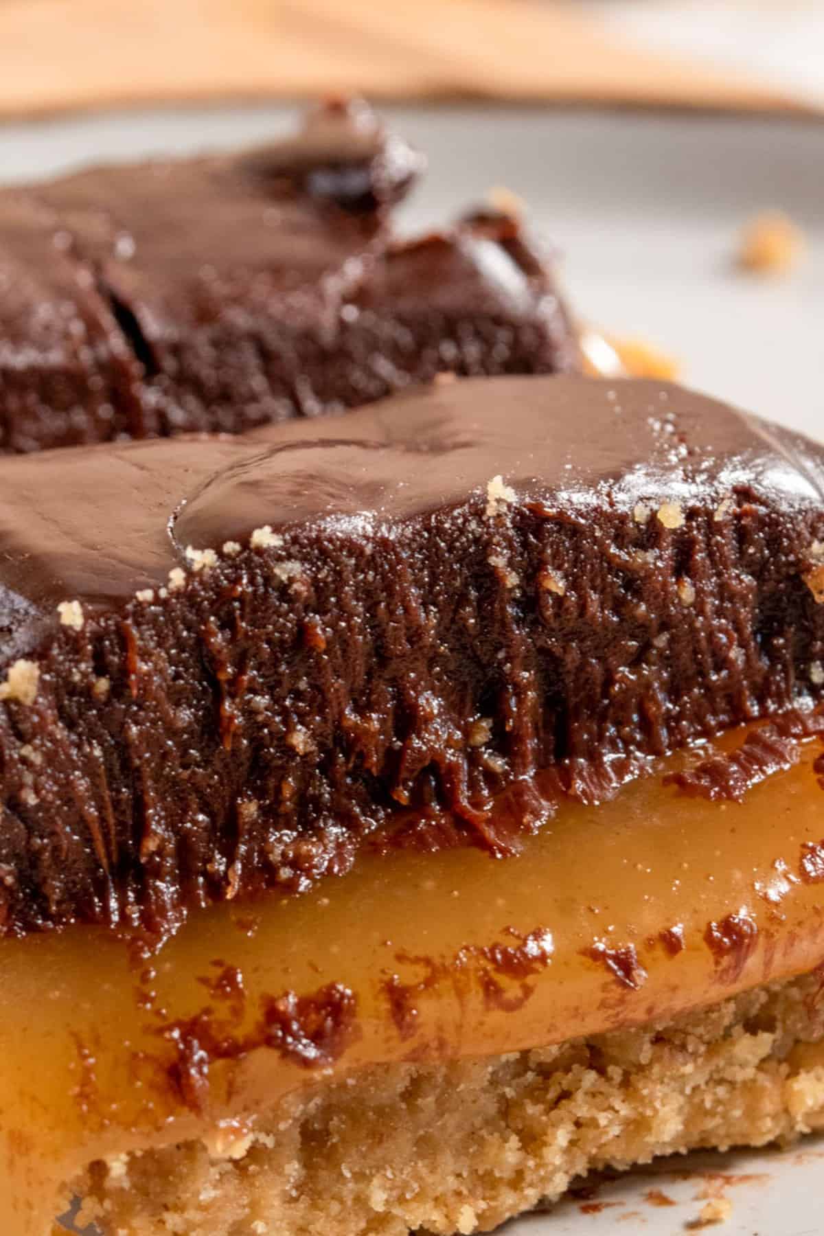 A super gooey vegan caramel slice on a brown-rimmed, white plate. The caramel is oozing out from the bar and the ganache topping is thick. 