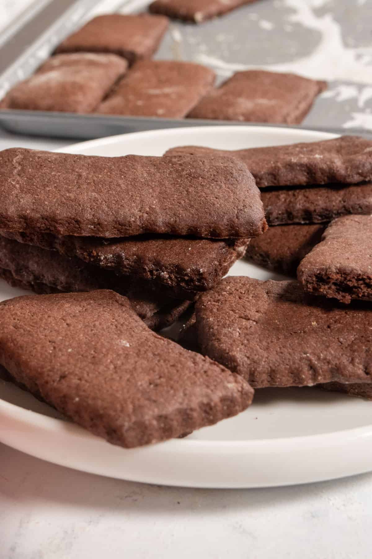 An untidy pile of vegan chocolate wafer cookies on a white plate. More sit on a baking tray in the background.