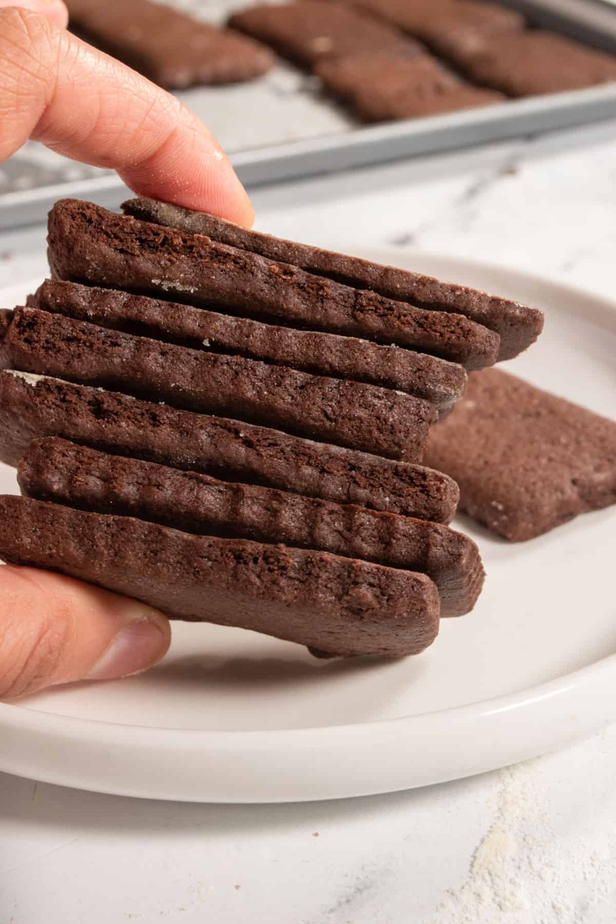 A hand holding a stack of chocolate cookies at an angle above a white saucepan.