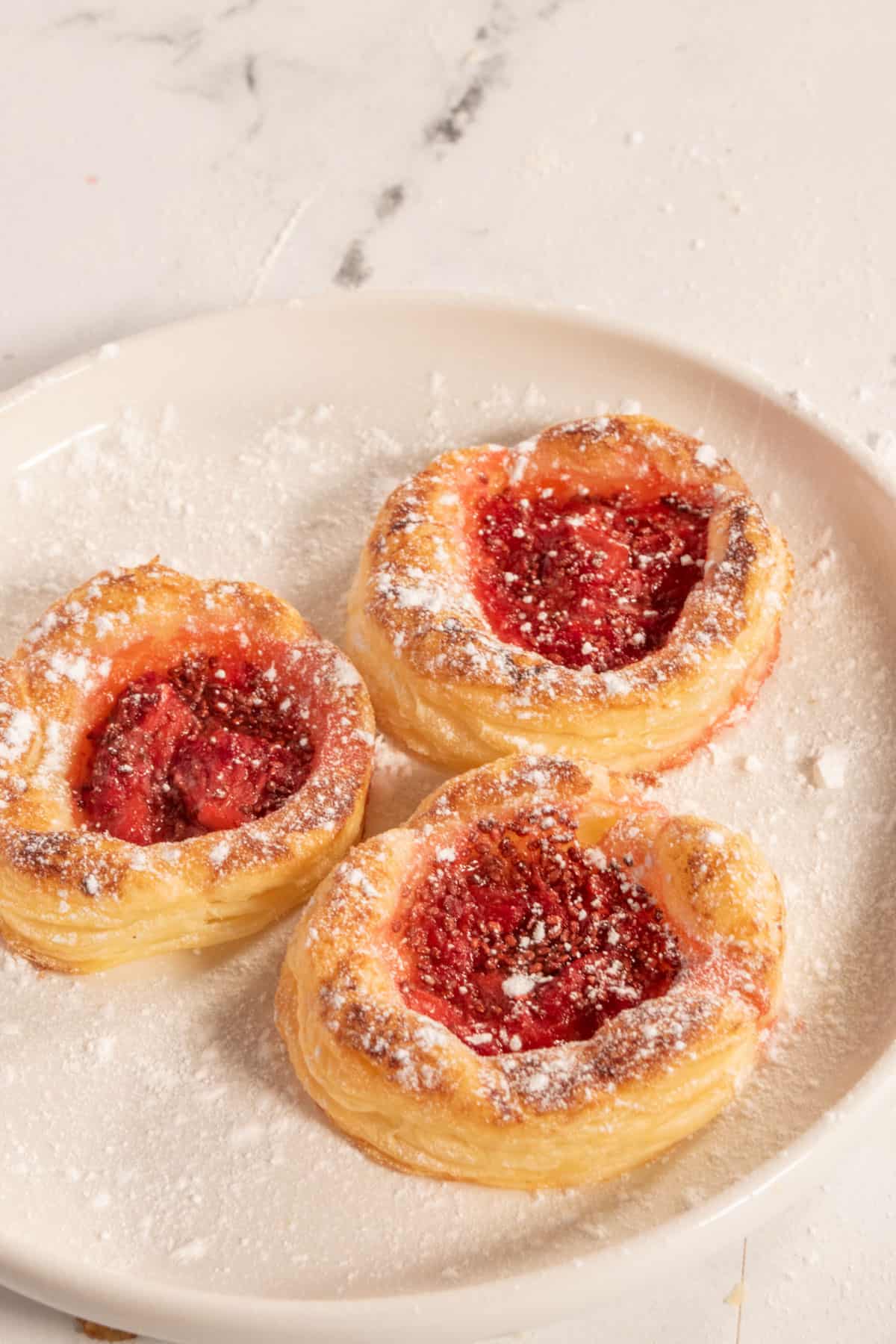 Three vegan Danish pastries on a white saucer, dusted with powdered sugar.