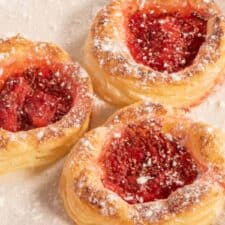 Three baked vegan Danish pastries on a white plate. They are golden brown and dusted with powdered sugar.