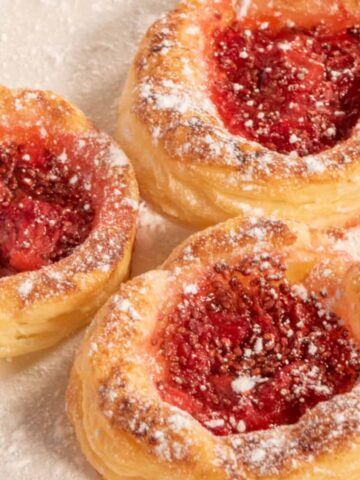 Three baked vegan Danish pastries on a white plate. They are golden brown and dusted with powdered sugar.