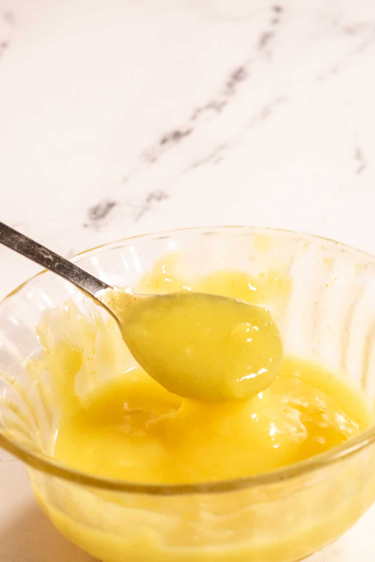 A small ramekin filled with bright yellow lemon curd. A small spoon leans on the side of it. 