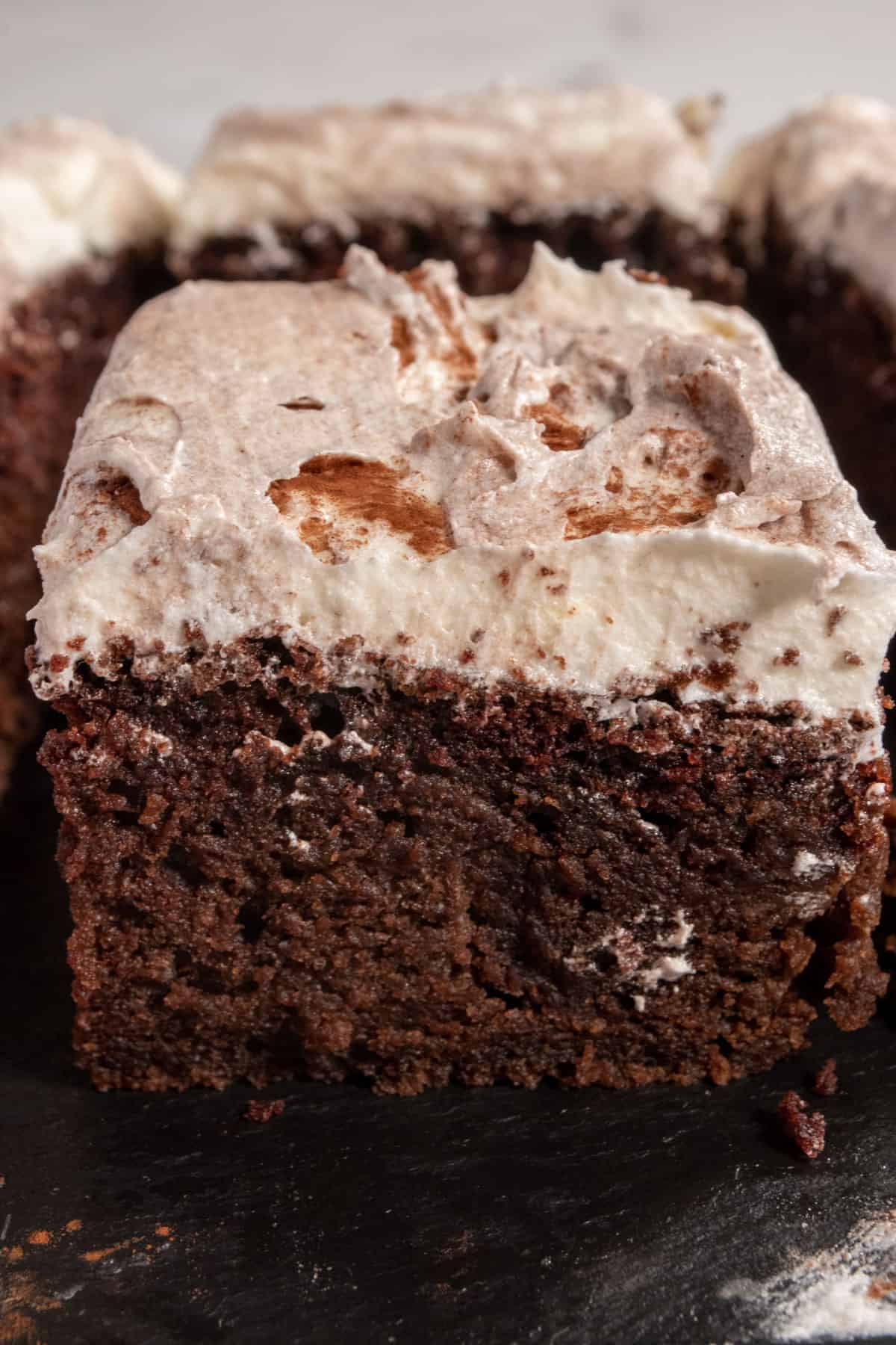 A tall slice of cake with a thick layer of frosting on top. The rest of the cake is in the background. 