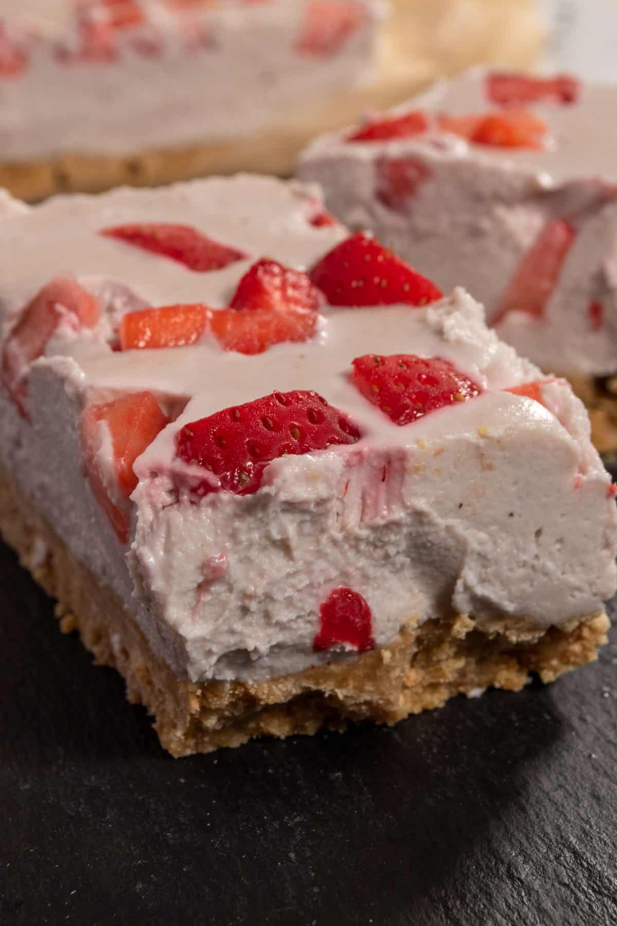 A creamy slice of vegan strawberry cheesecake on a slate platter. There are bright red fresh strawberries on top. 