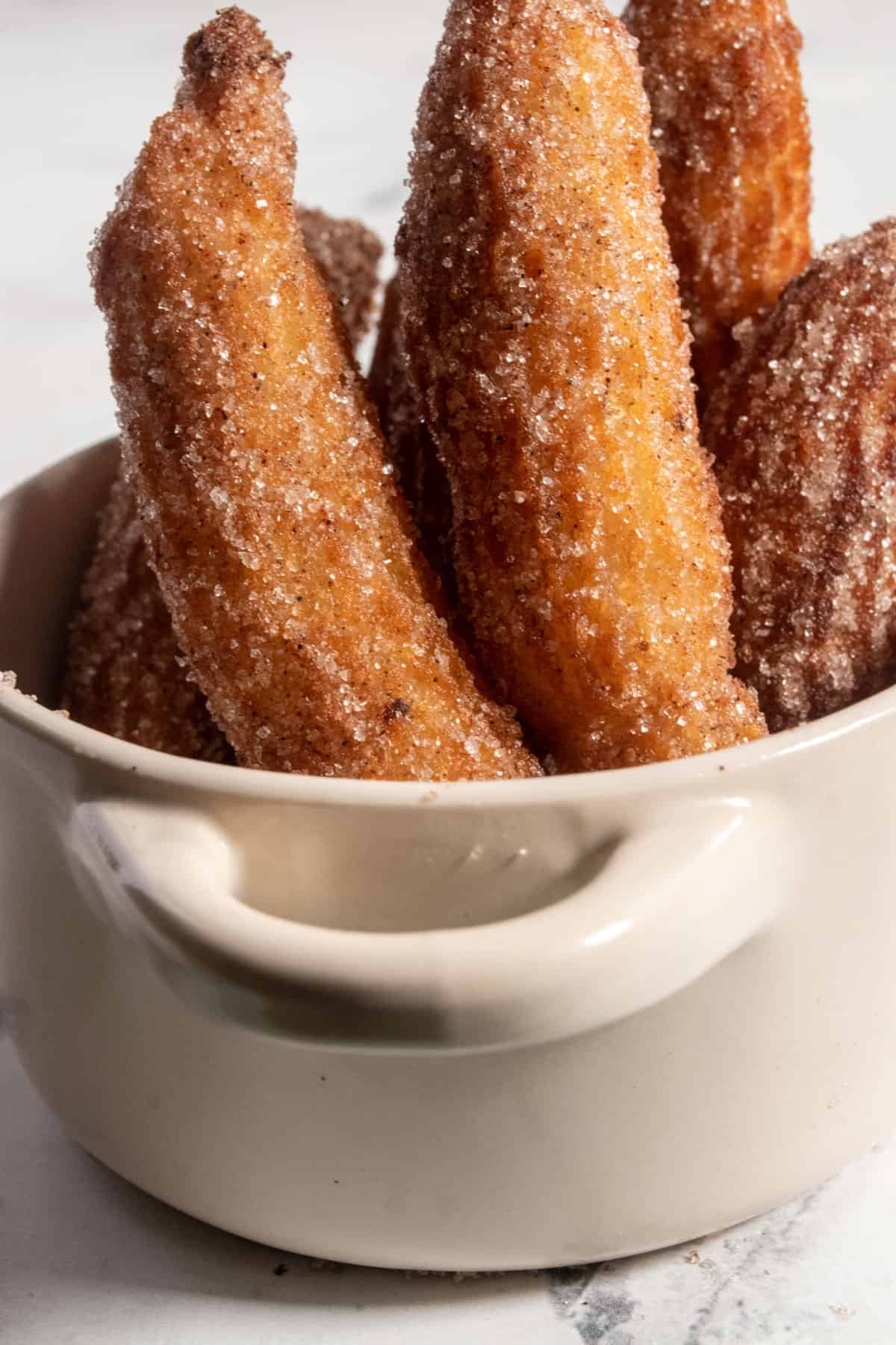 Six golden brown churros inside a white serving pot. They are covered in cinnamon sugar. 