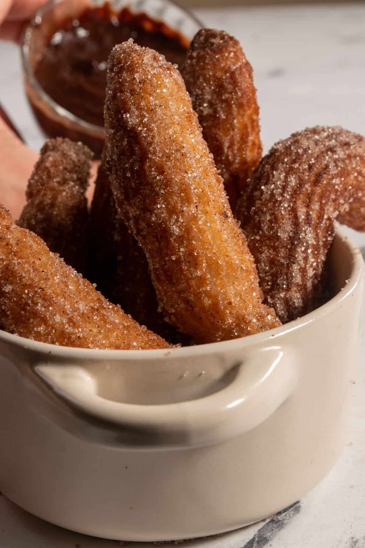 Golden brown, sugar-coated vegan churros inside a small ramekin. The chocolate dip is being held in the background. 