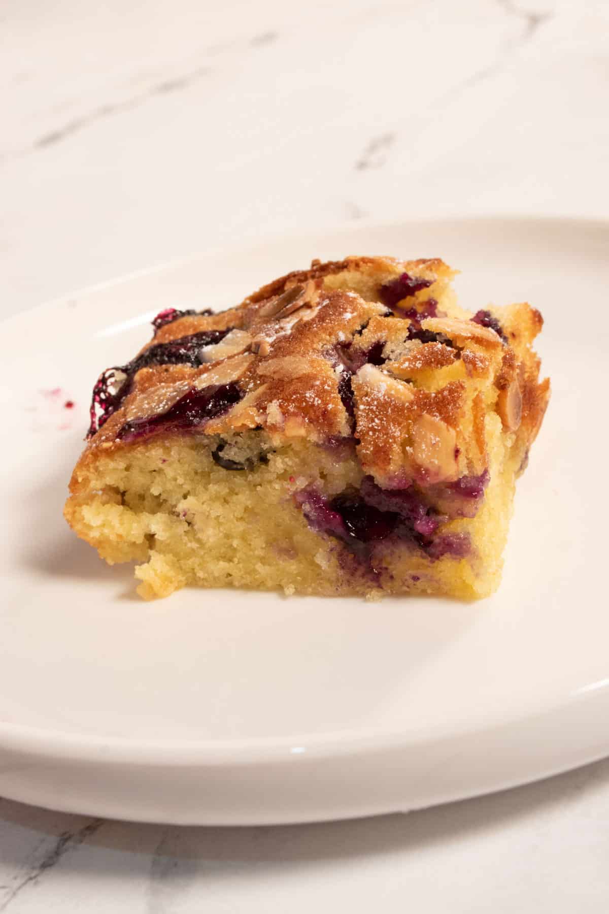 A small, super moist piece of blueberry cake on a white serving plate. It is packed with lots of blueberries and topped with toasty flaked almonds. 