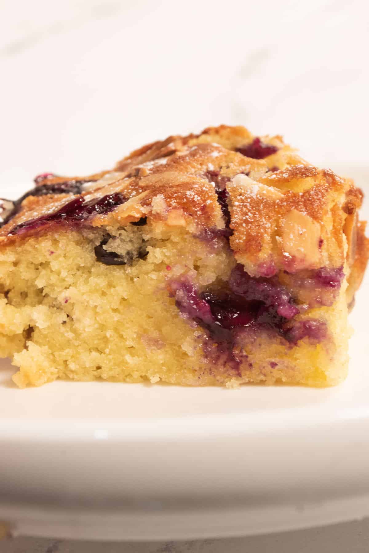 A golden piece of cake on a small, white saucer. The blueberries inside have burst and are dripping down the side of the cake. 