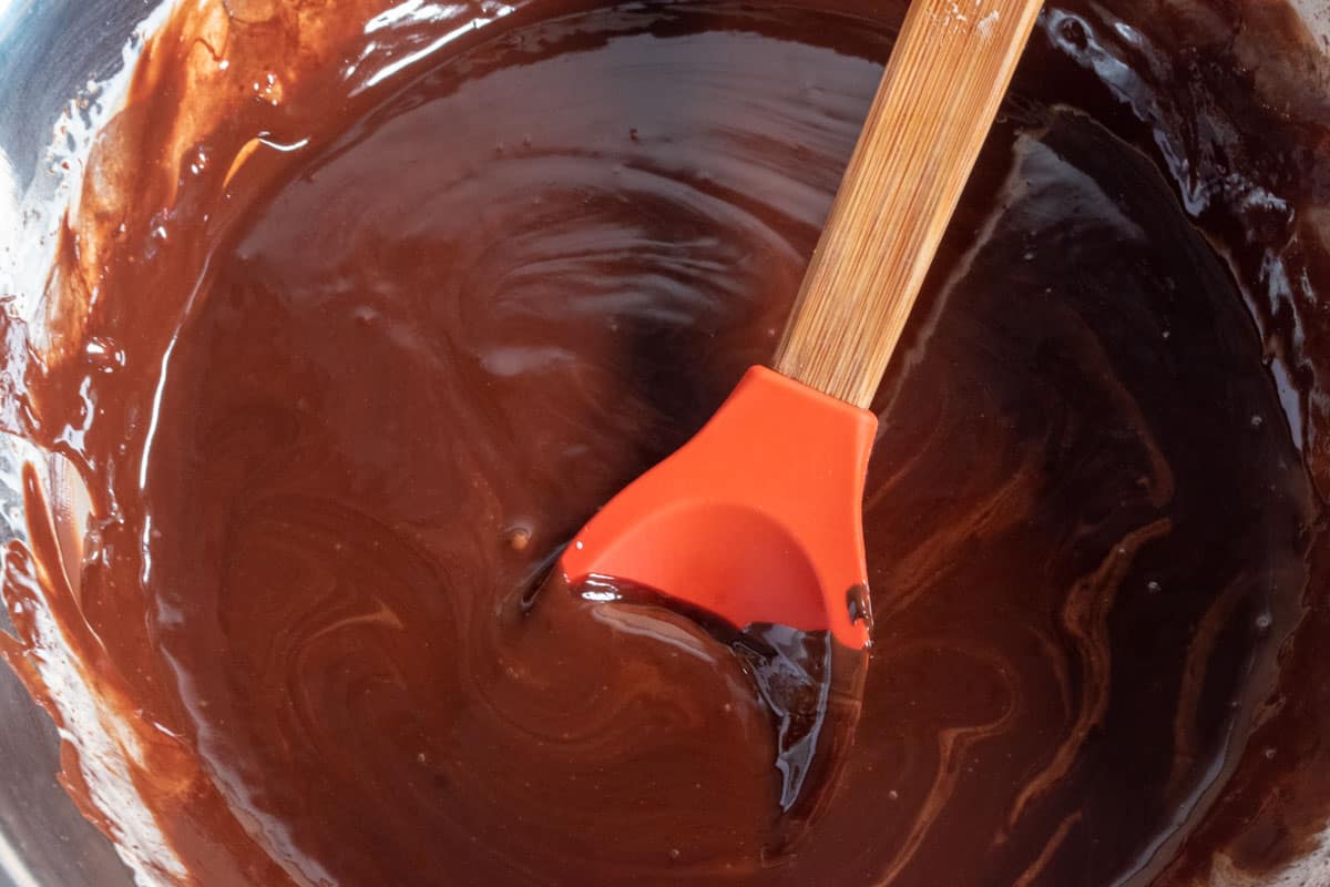 Chocolate being melted inside a large, metal mixing bowl. A red, silicone spoons sits inside the chocolate. 