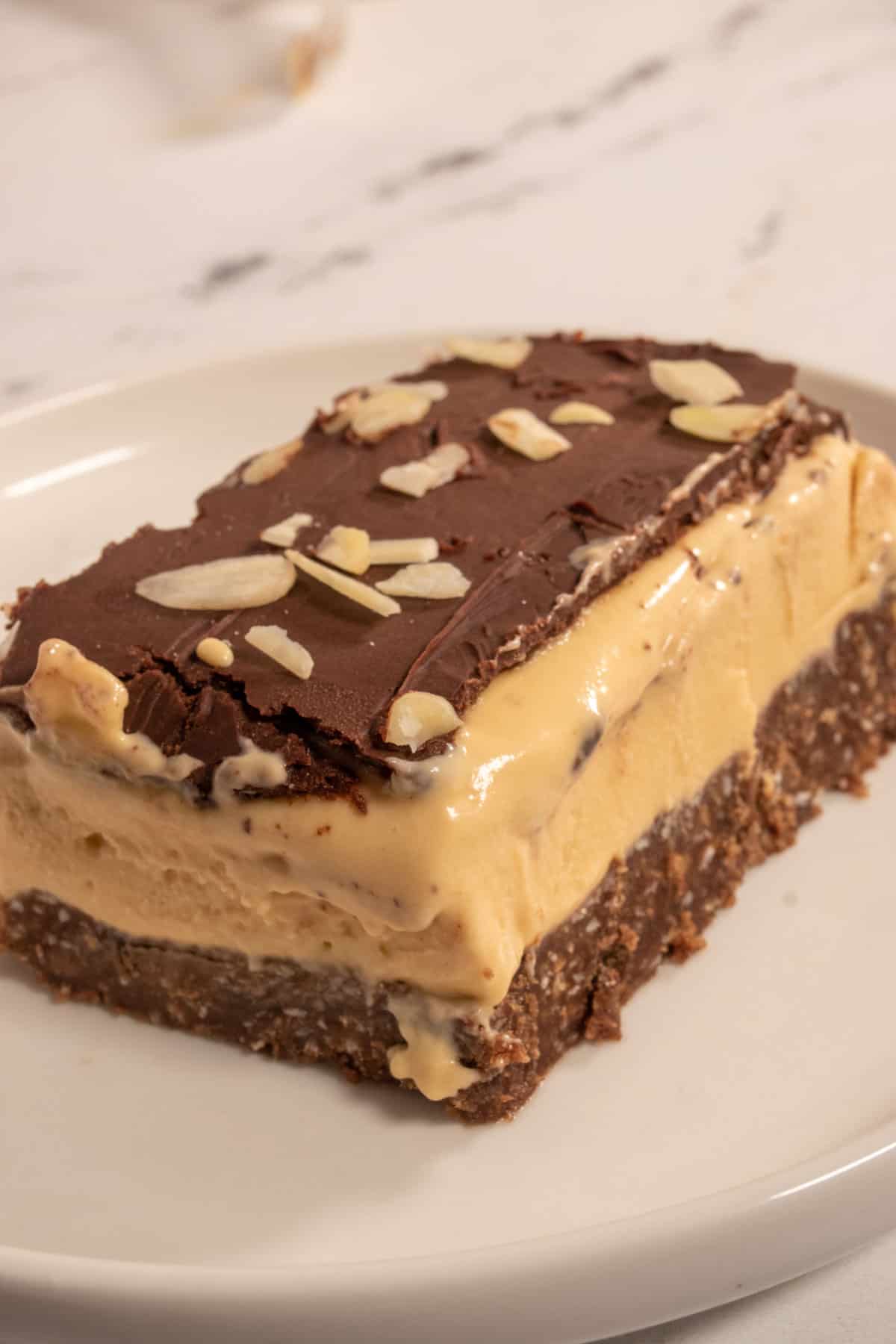 A single vegan Nanaimo bar custard slice on sitting on a white saucer. It is topped with flaked almonds and some of the filling is dripping down the sides. 