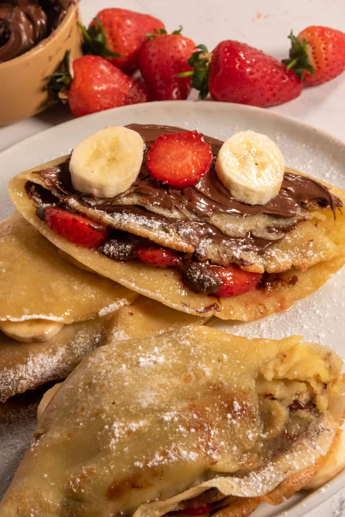 Many vegan oat milk crepes on a white serving plate, dusted with powdered sugar and filled and topped with bananas an strawberries.