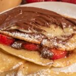 A chocolate covered vegan oat milk crepes on a white plate, filled with fresh fruit.