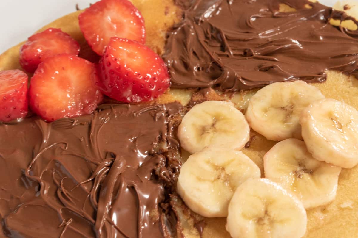 The crepe has been divided into quarters. Two quarters have been decorated with vegan chocolate hazelnut spread, one with sliced bananas and one with sliced strawberries. 