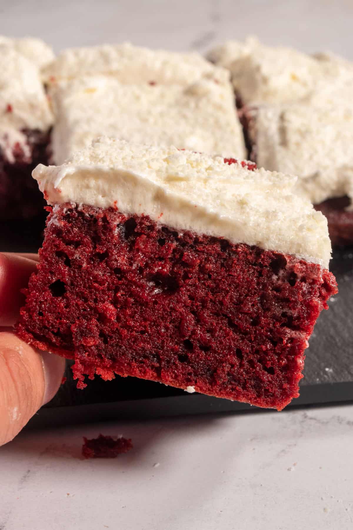 A hand holding a small piece of red velvet sheet cake, topped with white chocolate with other pieces in the background. 