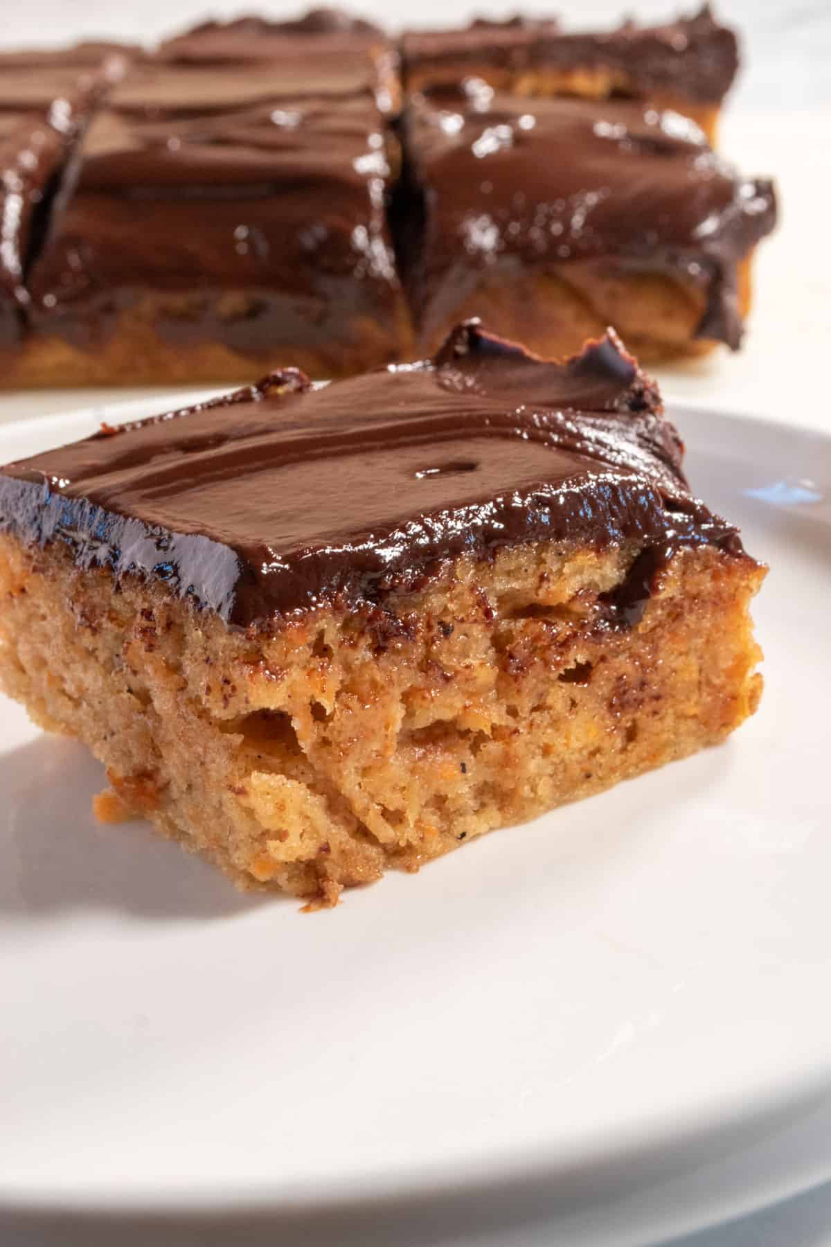 A super moist slice of carrot cake with a shiny layer of ganache on top on a white saucer. 