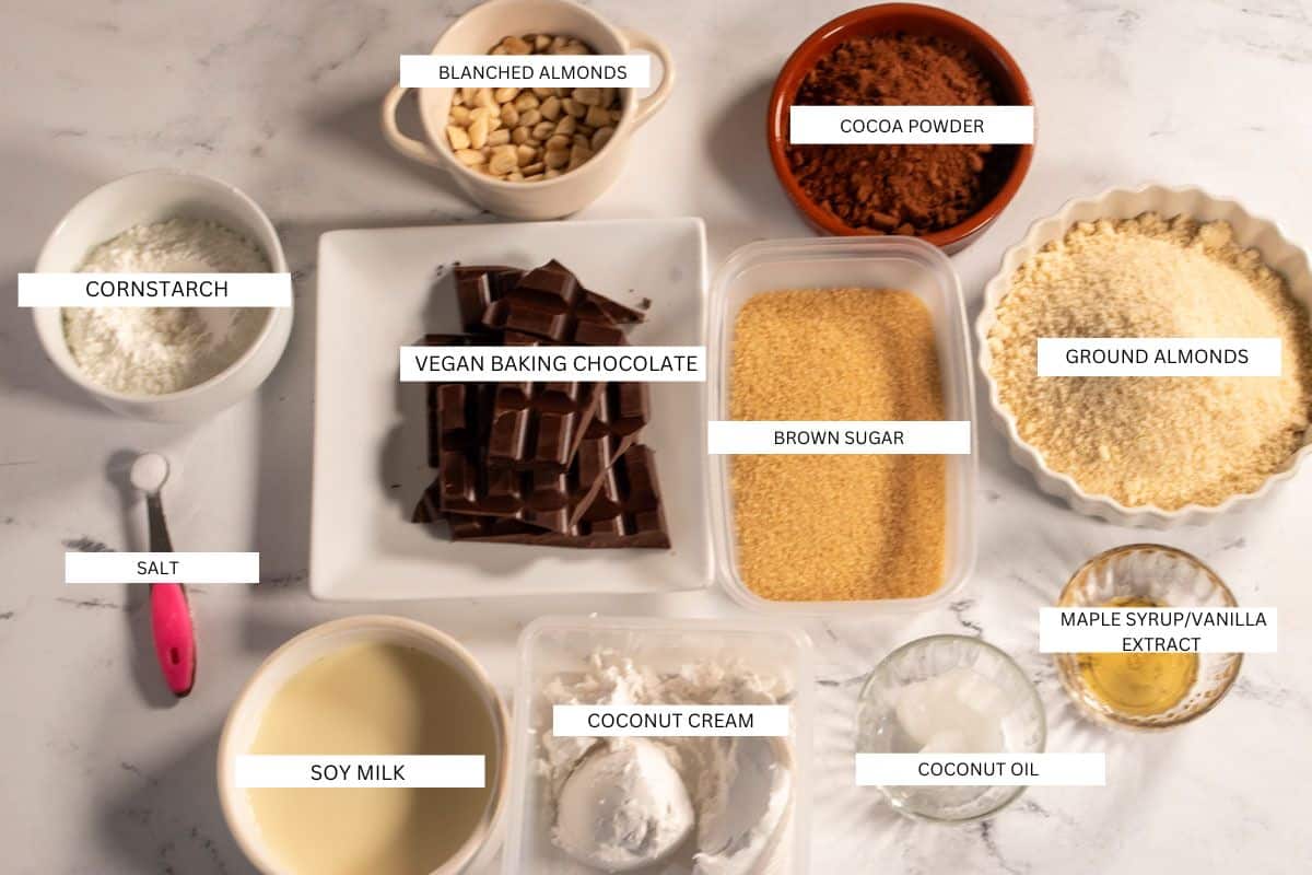 The ingredients that are required to make a chocolate vegan flourless almond cake, laid out neatly on a white marble background. 