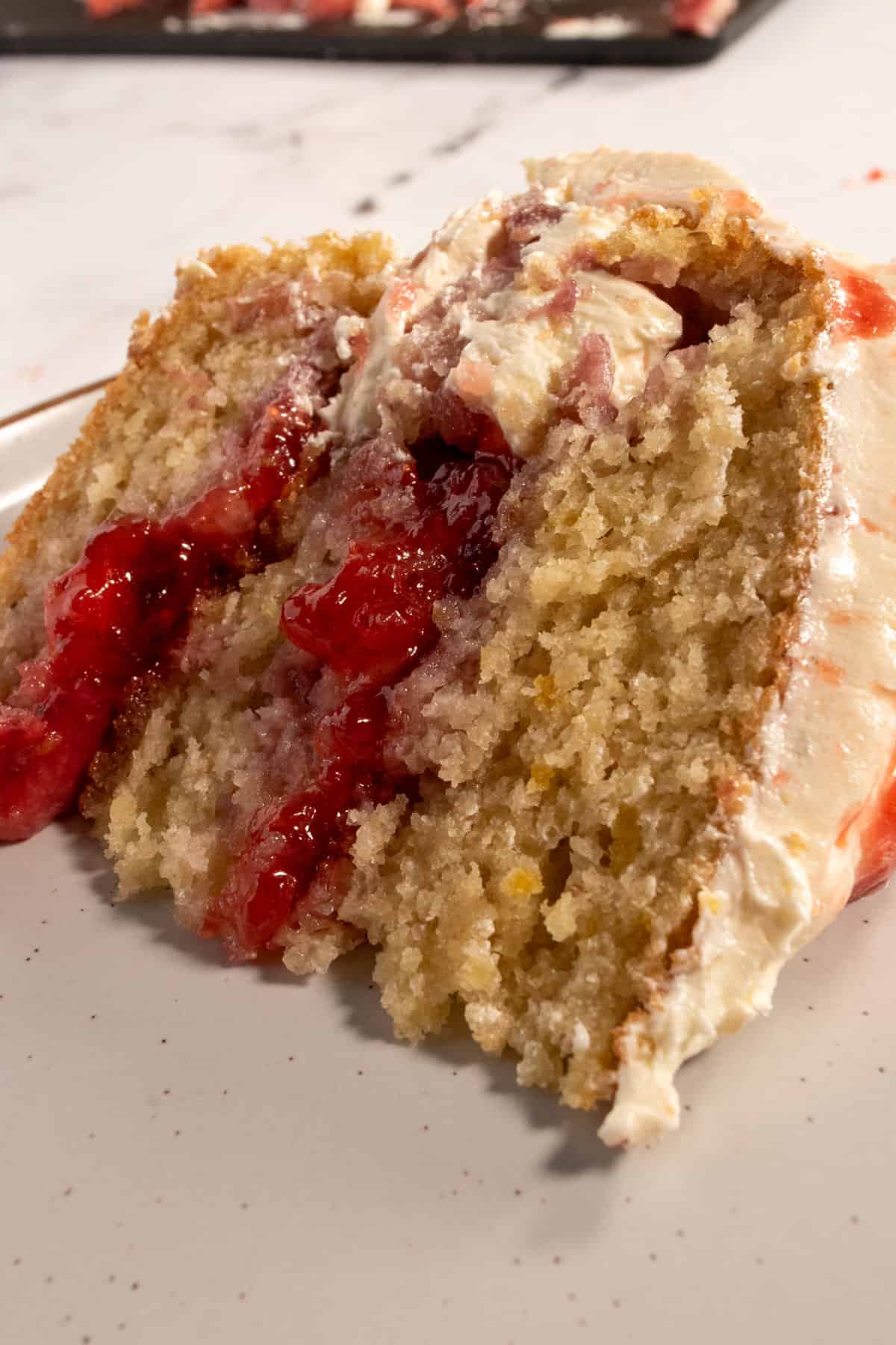 A large slice of vegan strawberry lemon cake on a white, brown-rimmed plate. There is lots of jam and frosting in the center.