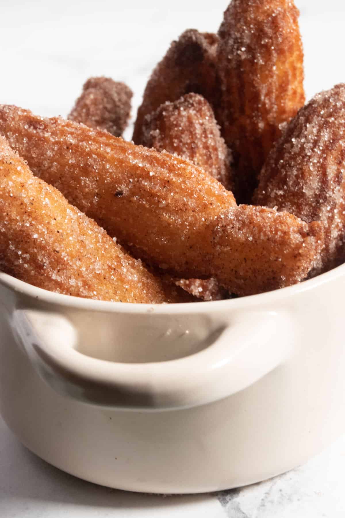 A small white pot filled with golden brown churros, which are covered in cinnamon sugar. 