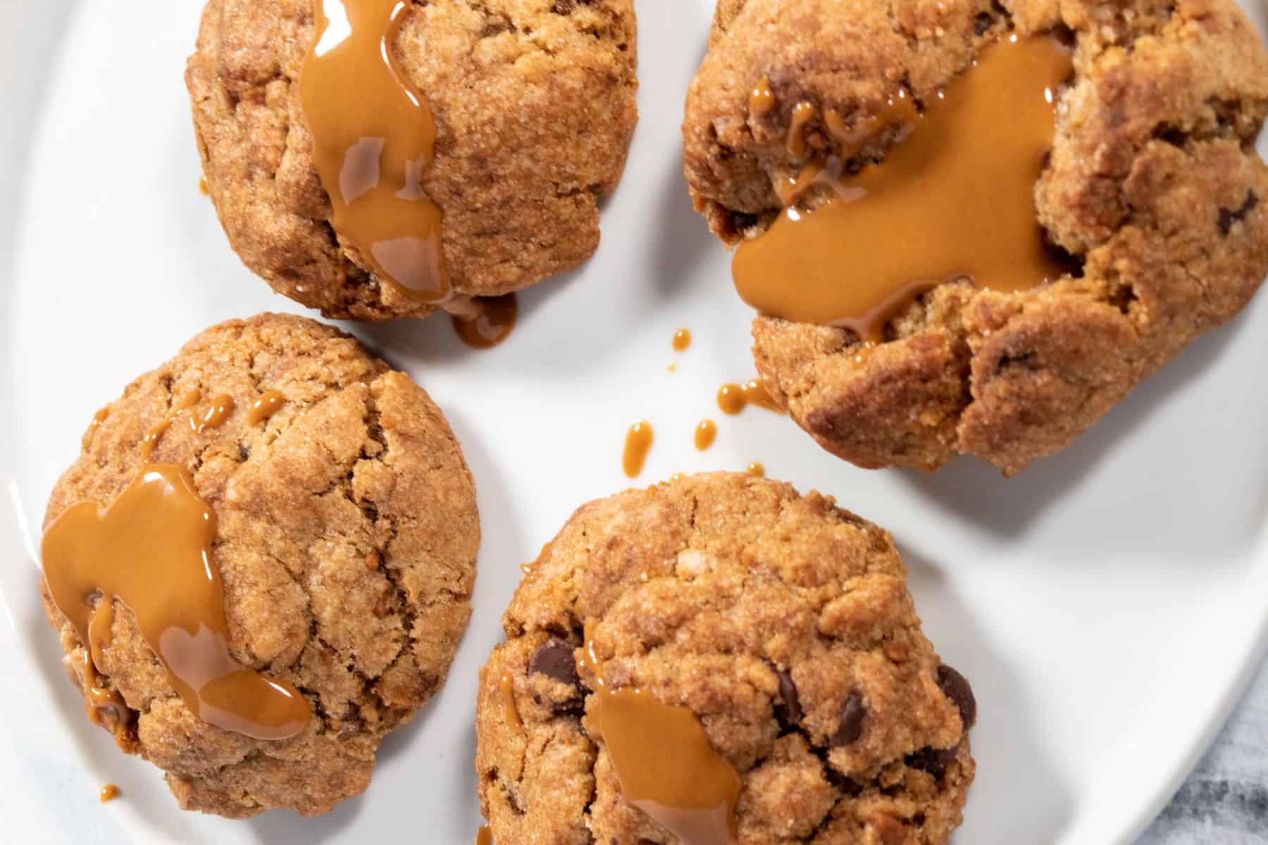 Four vegan Biscoff cookies on a larger-sized white plate. They have been decorated with melted Biscoff spread which drips down the sides of the cookies. 