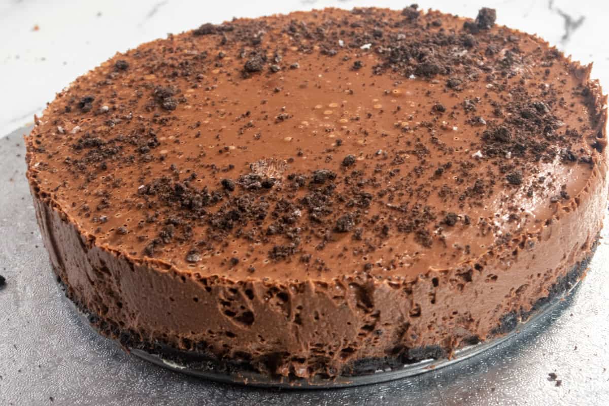 A full chocolate cheesecake on a metal platter. Oreos sprinkled on top. 