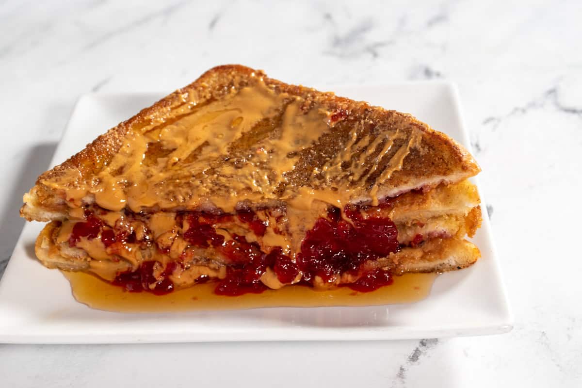 Chunky vegan peanut butter and jelly french toast on a small white saucer. Raspberry jelly and maple syrup is dripping down the sides.